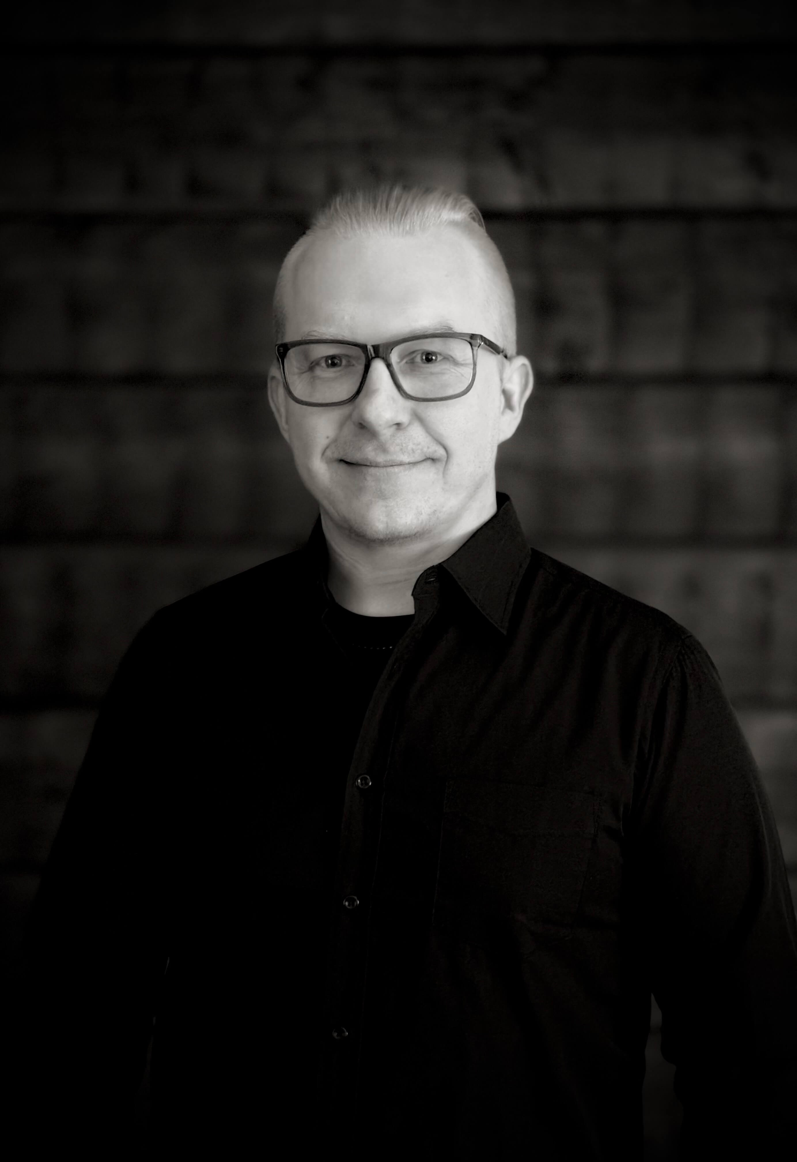 Mustavalkoinen muotokuva miehestä silmälasit päällä, musta paita ja puinen tausta.